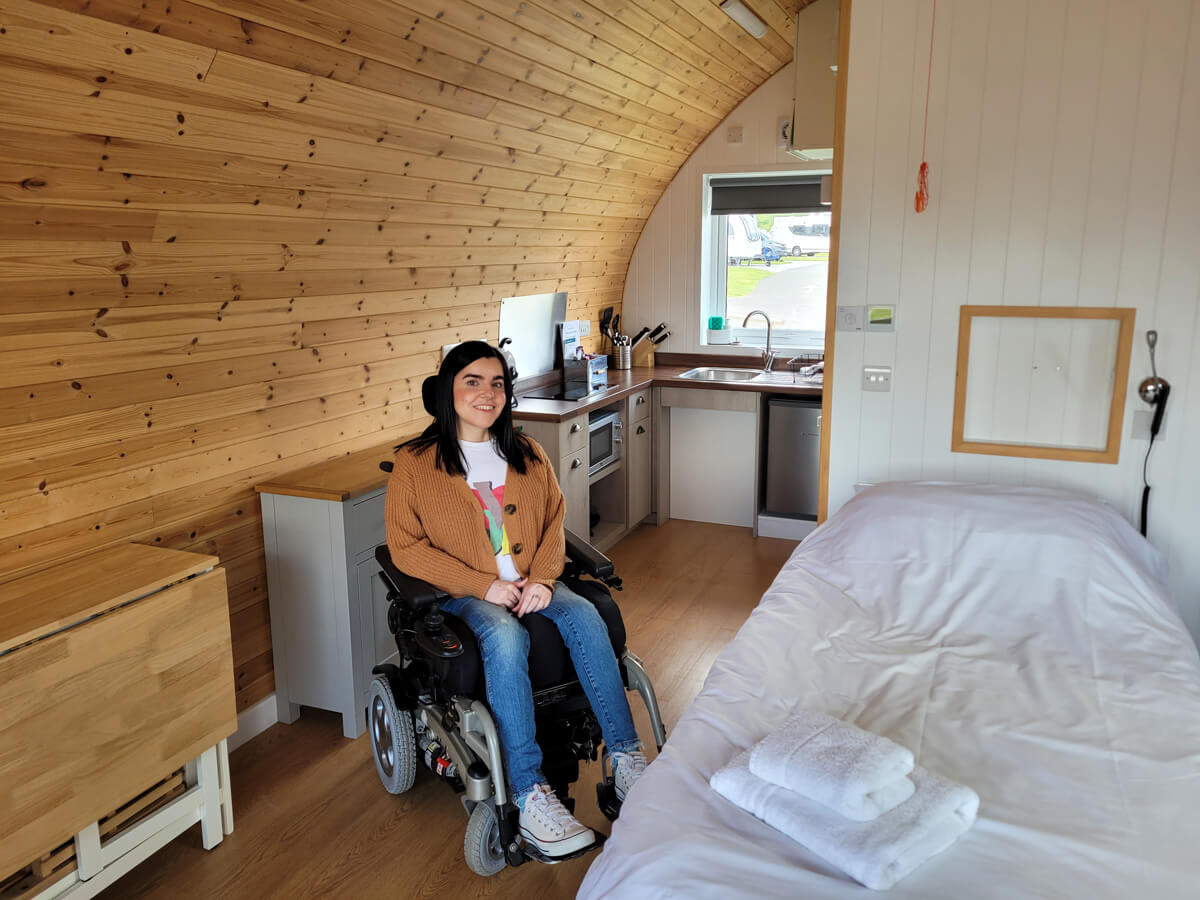 Emma sat inside the livingroom of the glamping pod. She is looking at the camera and smiling. The bed is on her left and the kitchen is in the background.