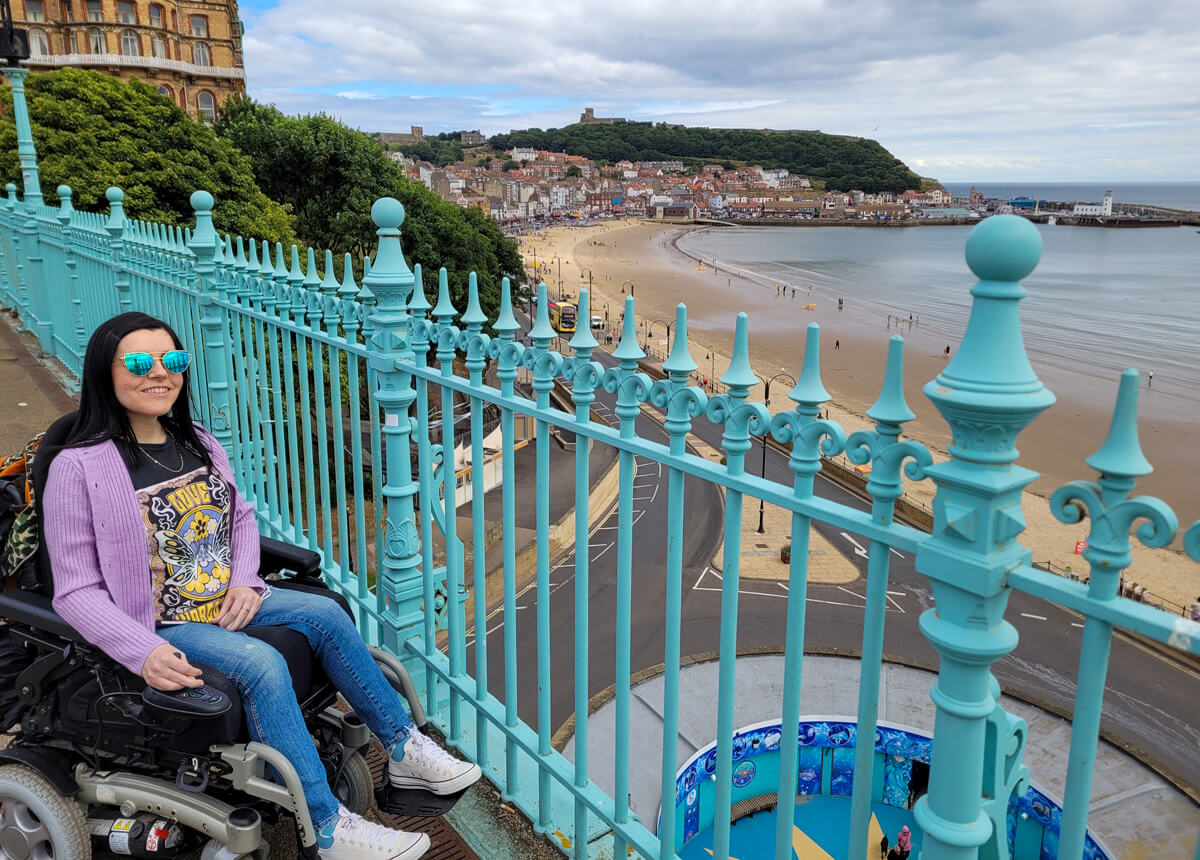 Emma sat in her wheelchair on Spa Bridge Scarborough. Emma is wearing a lilac cardigan and jeans. She is smiling and wearing sunglasses. There is a view of South Bay Beach.