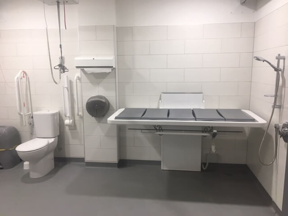 Interior of a Changing Places toilet showing the adult-sized changing bed, peninsular toilet and sink, ceiling-track hoist, grab rails and a shower.