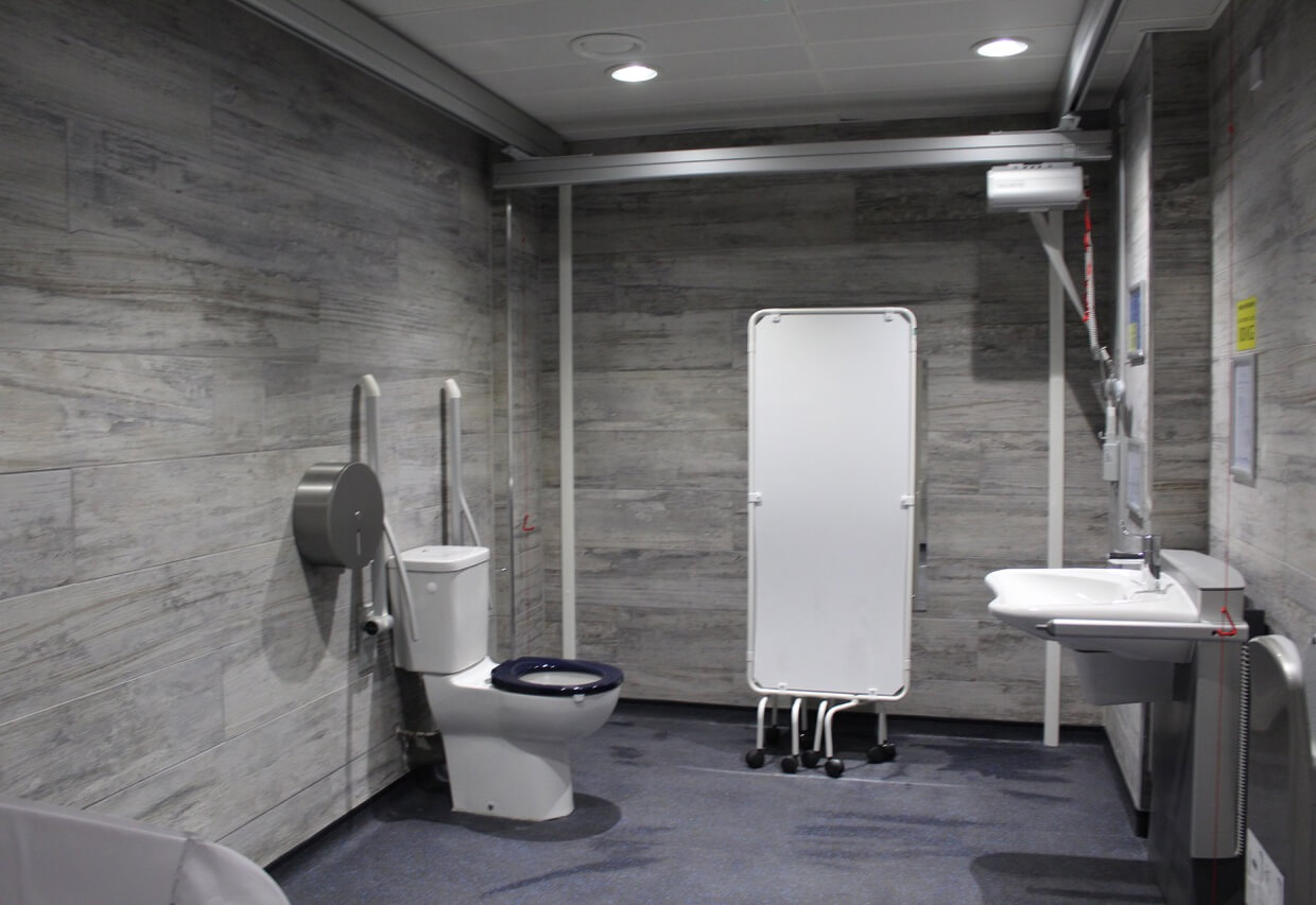 Interior of a Changing Places toilet showing the adult-sized changing bed, peninsular toilet and sink, ceiling-track hoist and grab rails.