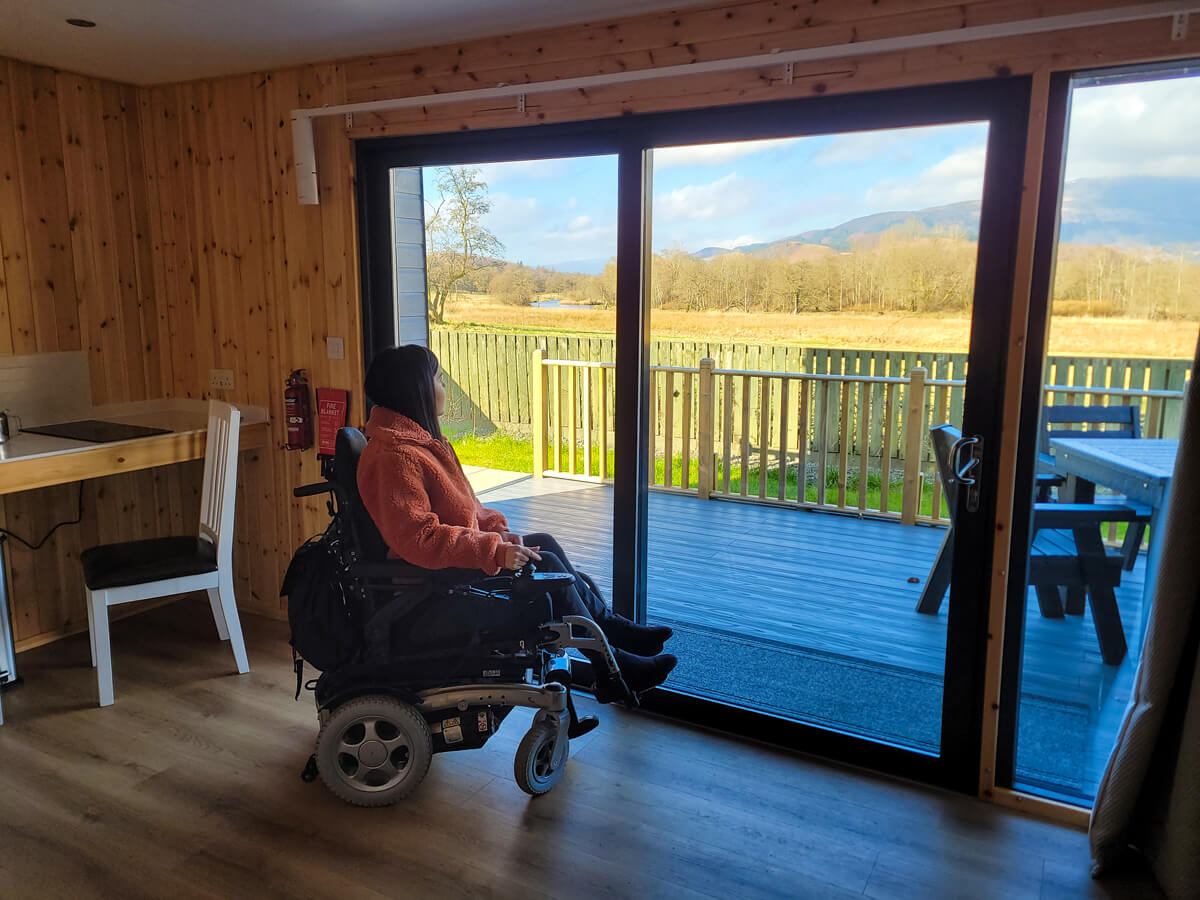 Emma sat in her powered wheelchair at a ceiling to floor window. She is looking out of the window at the stunning view of the mountains.