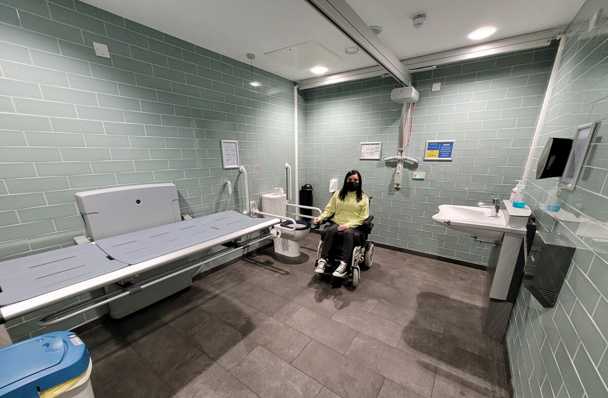Emma, a white female powerchair user with dark shoulder length hair is is a Changing Places toilet. The walls are mint green brick tiles and the floor is grey slate tiles. There is adult-sized changing bed, peninsular toilet and sink, ceiling-track hoist, grab rails and a shower.