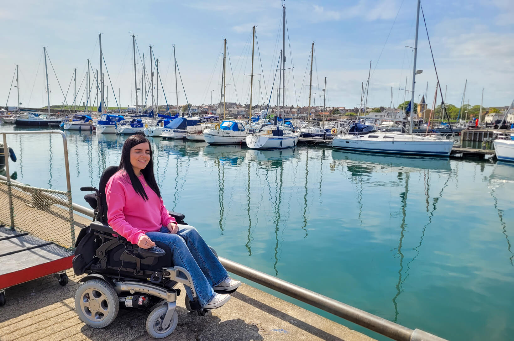 Emma sat in her wheelchair at Anstruther Harbour. The water is turquoise colour and there are numerous boats behind her. Emma has long black hair and wearing a pink sweatshirt.