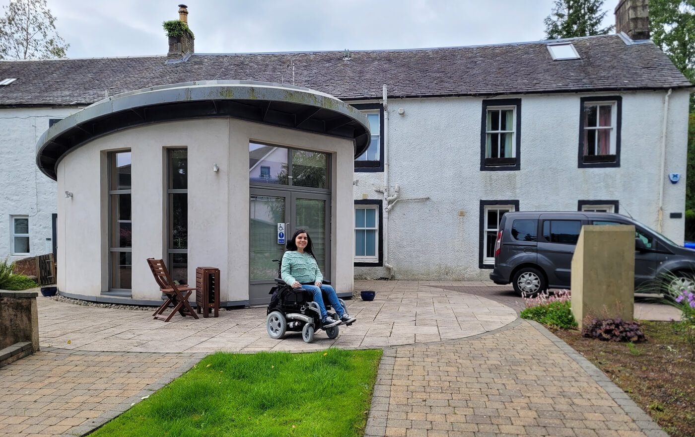 Emma in her wheelchair sat in outside in front of Clober Farm, a wheelchair accessible cottage in Scotland. Emma is wearing a mint green cardigan and blue jeans. Her grey wheelchair accessible vehicle is parked on the right.