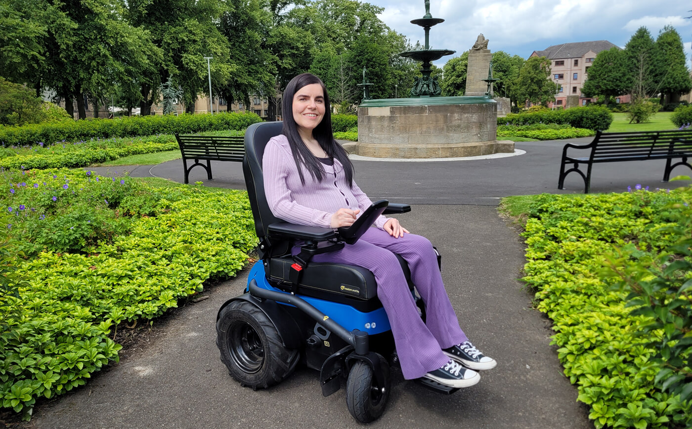 Emma, a wheelchair user sitting in a blue and black Freedom One Life Series 5 Powerchair in a park. She is wearing purple flared leggings and a purple cardigan.