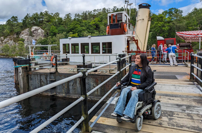 Emma sits in her wheelchair wearing a black jacket, multicoloured striped jumper, blue flared jeans and black converse. Her long dark hair is styled down. She is on the wooden bridge with Steamship Sir Walter Scott stationed behind her. Emma is looking to the side and smiling.