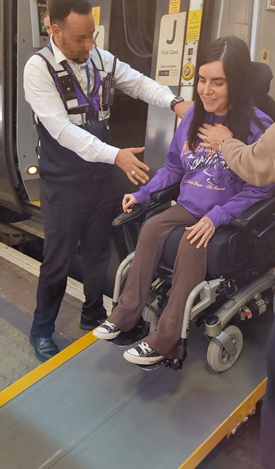 Emma, a power wheelchair user is wearing a purple sweatshirt and brown flared leggings. She is exiting the train via a wheelchair ramp at London Euston station. She is smiling. The Network Rail customer service representative is standing next to Emma.