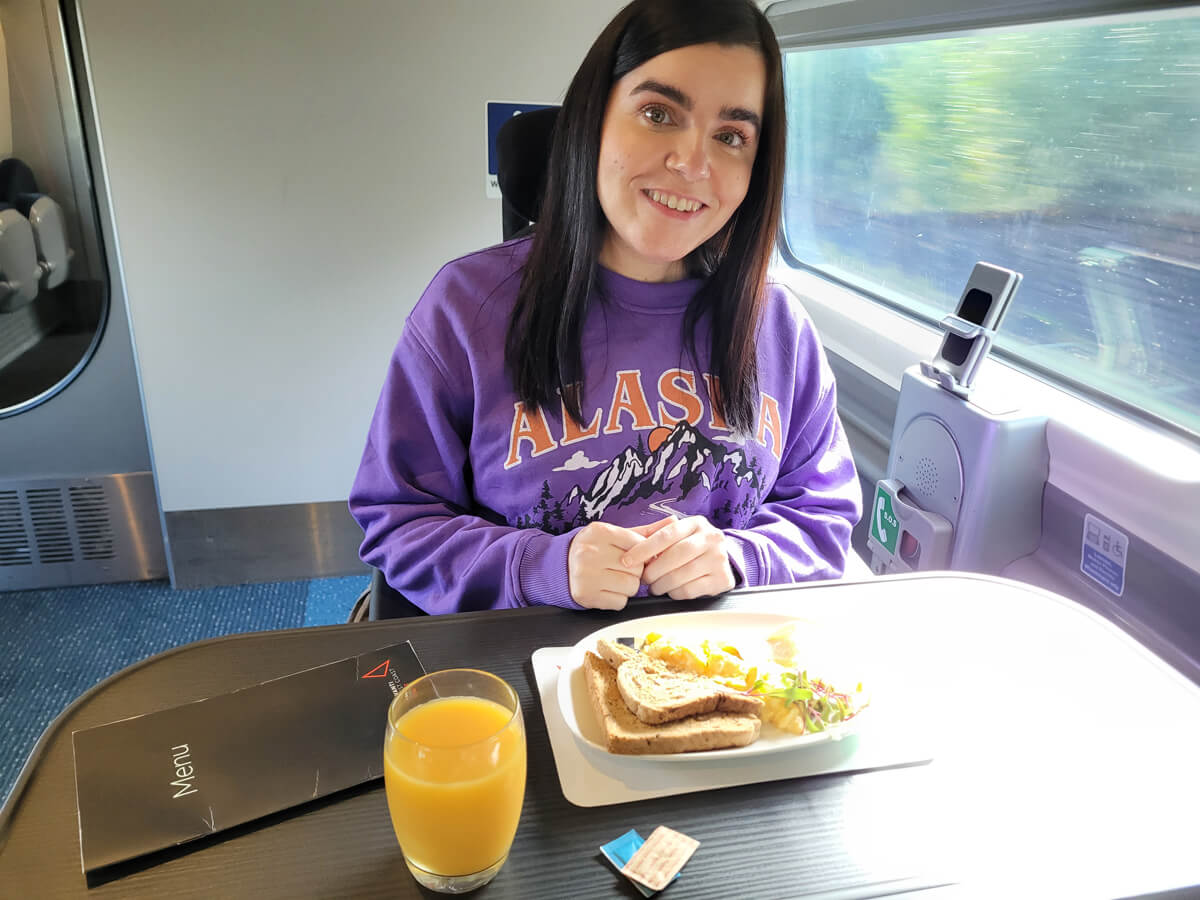 Emma is wearing a purple Alaska sweatshirt. She is in the wheelchair space on Avanti West Coast train. She has a plate with toast and scrambled eggs and a glass of orange juice.