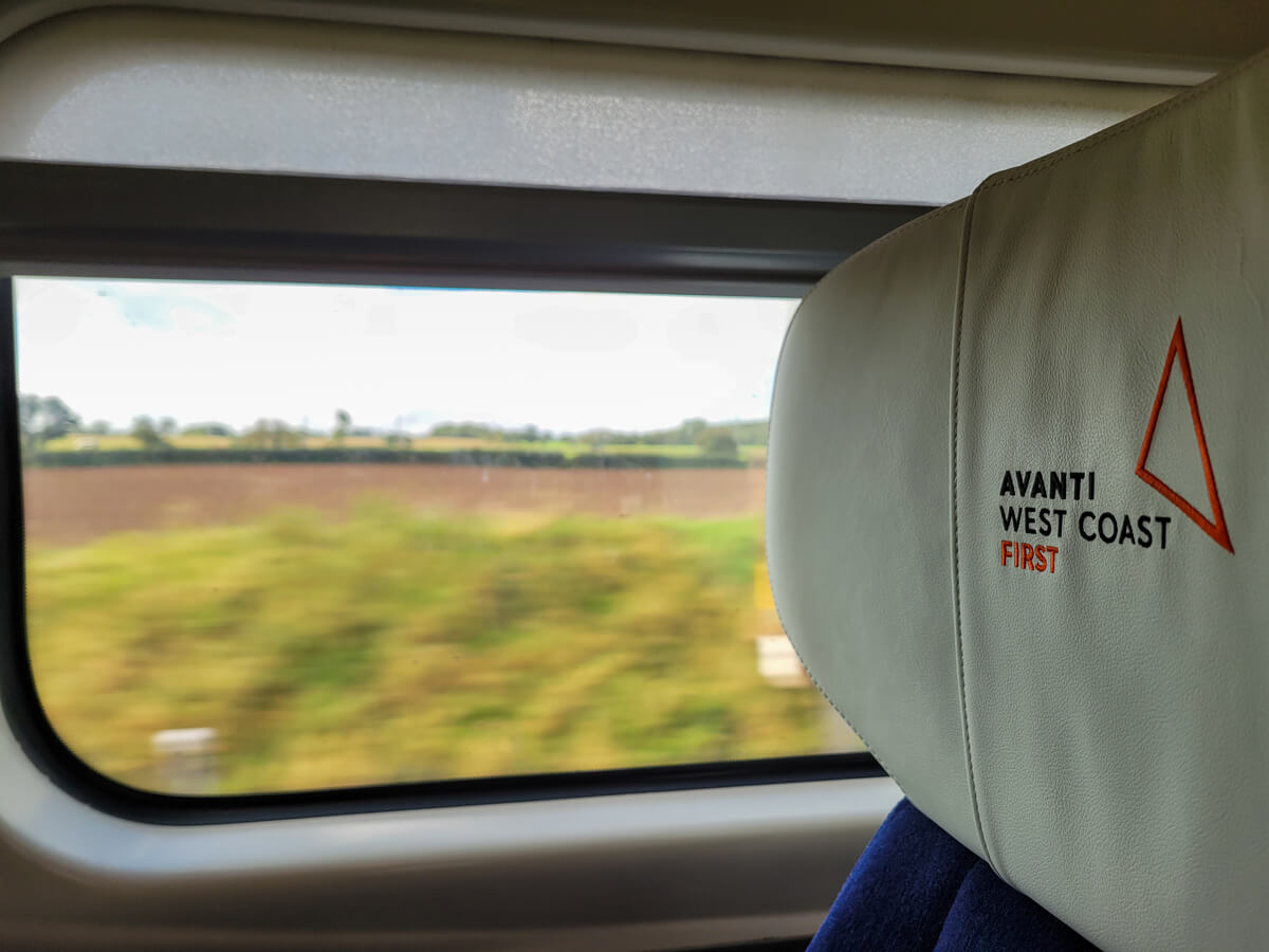 A close up shot of the train seat showing the Avanti West Coast First headrest with a view of the countryside out of the window.