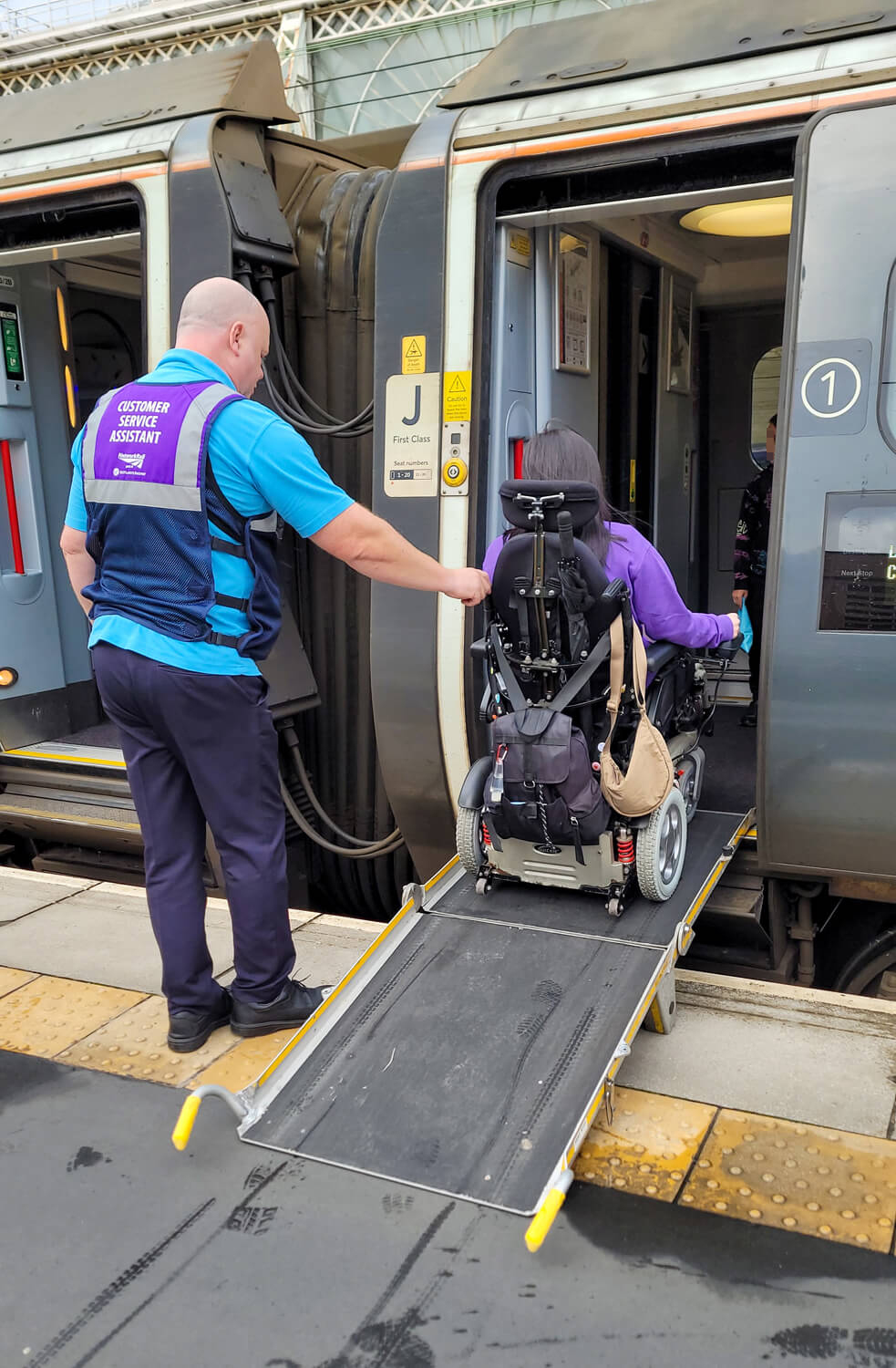 Emma driving her power wheelchair up a boarding ramp into an Avanti West Coast train. The Network Rail customer service representative is standing behind Emma holding on to the back of her chair.