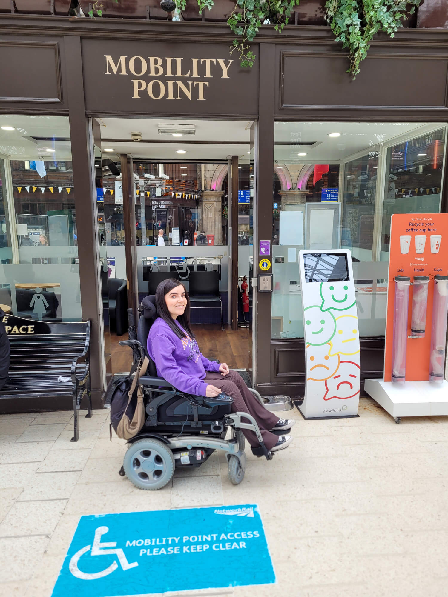 Emma, a power wheelchair user is wearing a purple sweatshirt and brown flared leggings. She is sat next to the Mobility Point office inside Glasgow Central Station.
