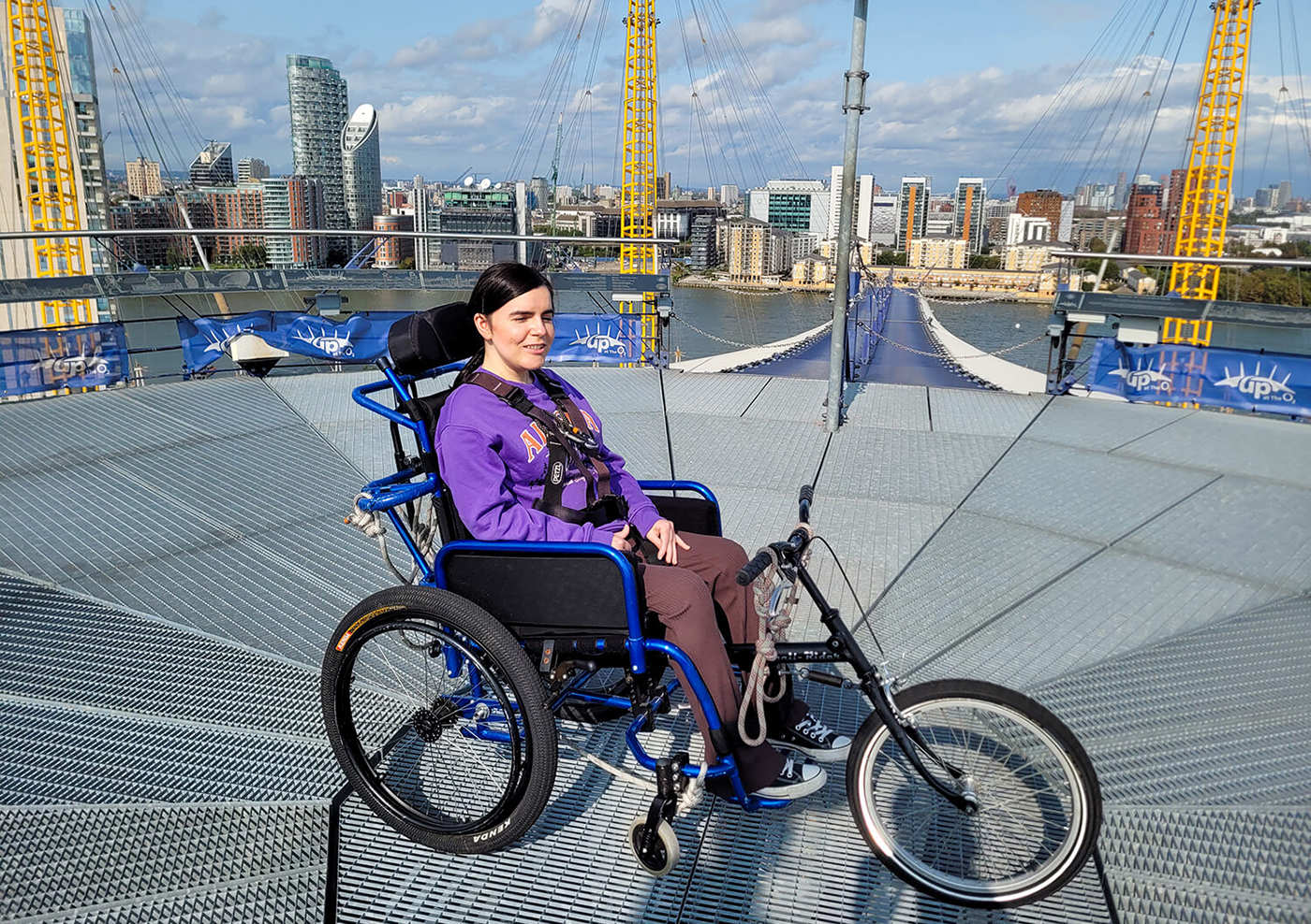 Emma is sitting in a manual wheelchair provided by Up at the O2 for the purpose of her O2 wheelchair climb. She is on the summit of the O2 roof with a spectacular view around her. Emma is wearing a purple sweatshirt and brown flared leggings. Emma is smiling.