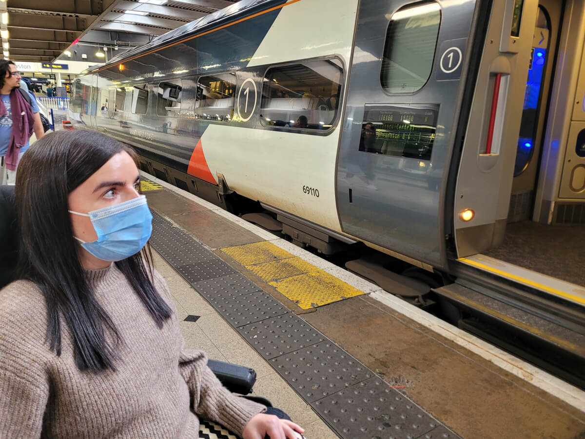 Emma and female wheelchair is wearing a face mask. She is sat next to an Avanti West Coast train waiting to board.