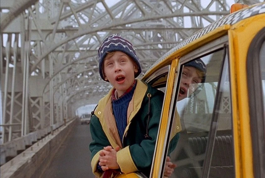 A scene from Home Alone 2: Lost in New York where Kevin is leaning out of a yellow cab driving over a New York bridge.