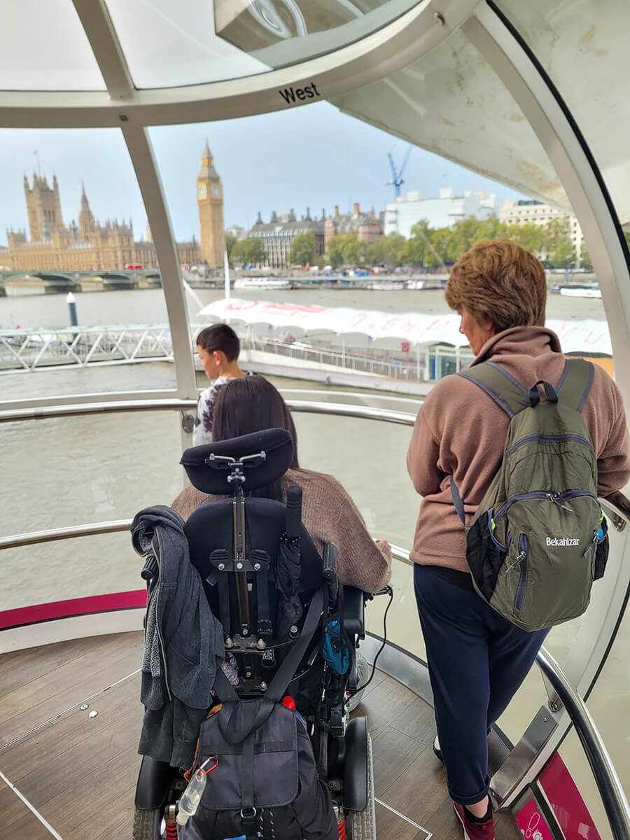 Emma sat in her power wheelchair inside the London Eye next to her Mum and nephew. They are looking out the window at the view of the river Thames and Westminster.