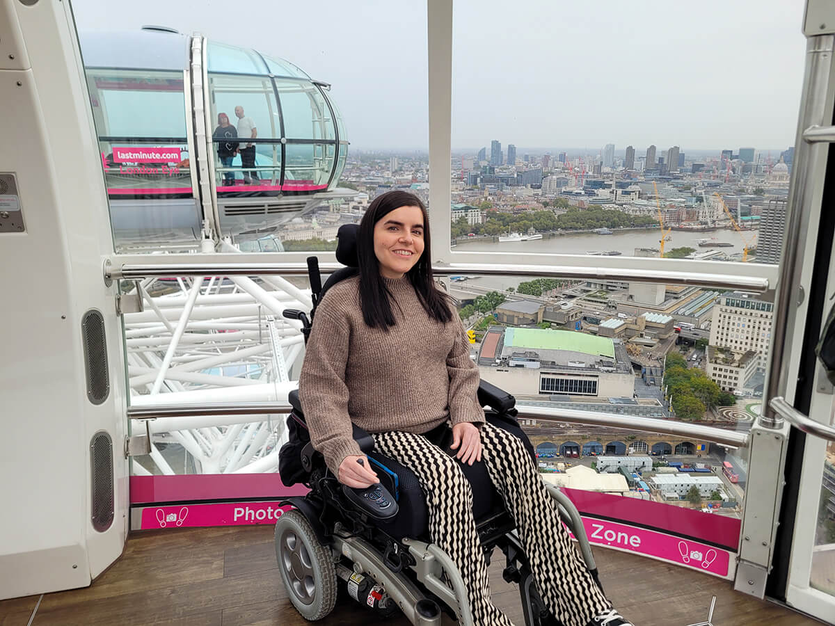 Emma sat in her power wheelchair inside the London Eye. Behind Emma is a view of the river Thames and tall buildings. Emma is wearing a light brown jumper and black and beige trousers.