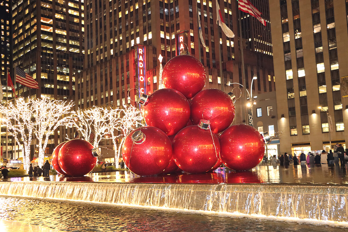 a group of red Christmas baubles sitting on top of a fountain. The Radio City Music Hall is behind the baubles.