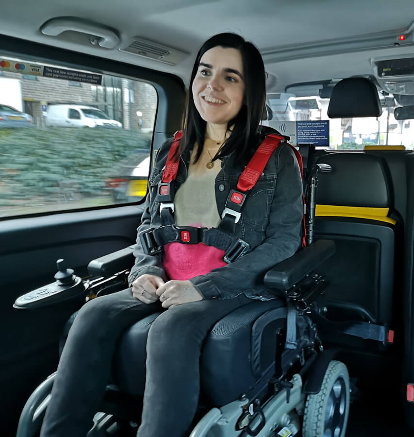 Emma sitting in her wheelchair wearing a harness seat belt in the back of a taxi.