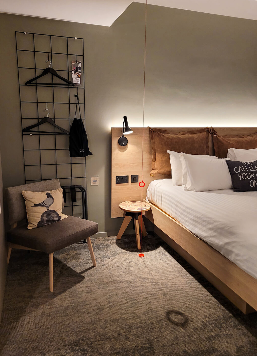 A photo showing the side of the bed, bedside table, emergency red cord, open wardrobe and chair in the accessible bathroom at the Moxy Manchester City hotel.