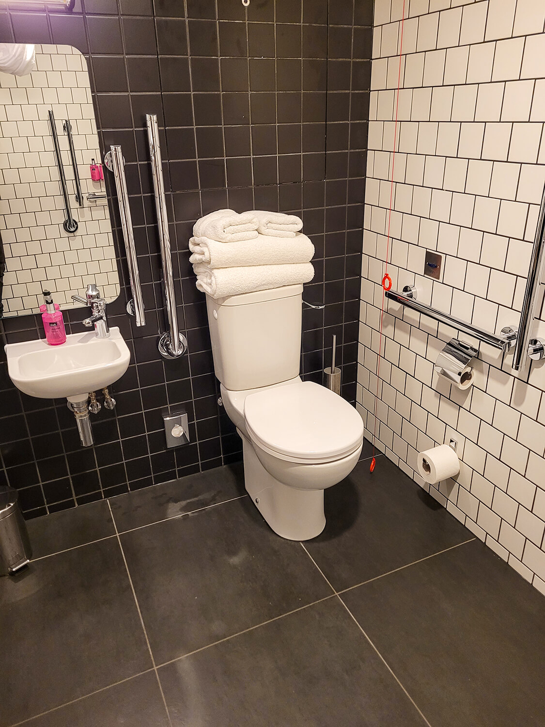 The toilet with grab rails and emergency red cord in the accessible bathroom at the Moxy Manchester City hotel.
