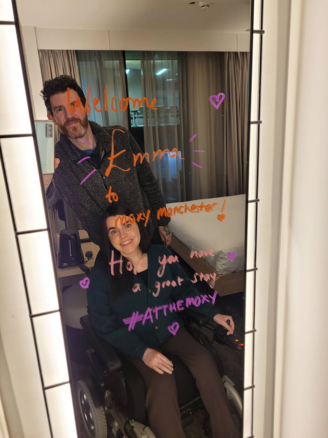 A mirror selfie of Emma and Allan. There is a handwritten message on the mirror saying "Welcome Emma to Moxy Manchester. Hope you have a great stay #AtTheMoxy