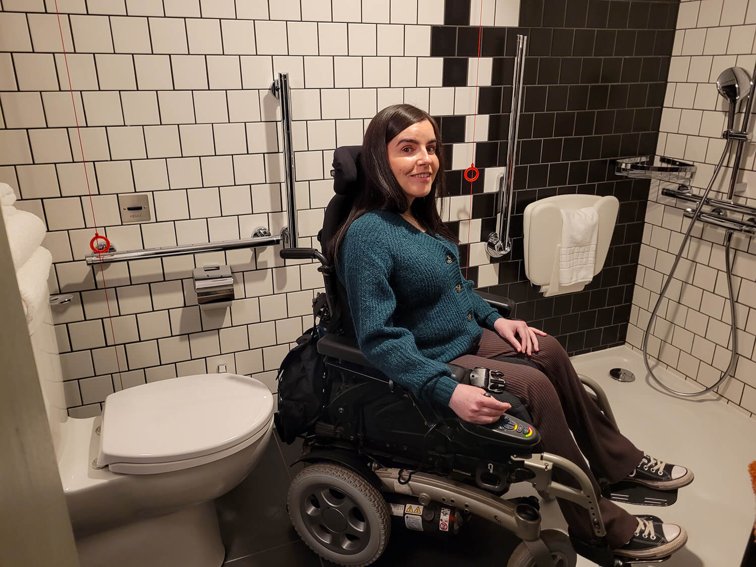 Emma sat in her wheelchair in the accessible bathroom at the Moxy Manchester City hotel. She is wearing a dark green cardigan, brown flare trousers and has long black hair.