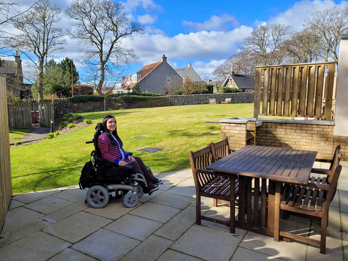 Emma in her wheelchair sat in the garden at Homelands . It is beautifully landscaped. Emma is sat next to a four seat table. She is smiling at the camera.