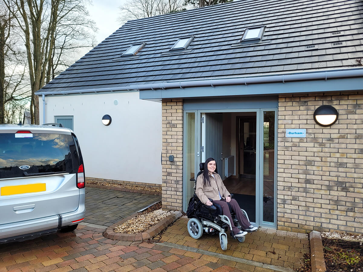 Emma in her wheelchair sat in outside in front of the Durham lodge at Homelands Fife, a wheelchair accessible lodge in Scotland. Emma is wearing a beige jacket and brown trousers. Her silver wheelchair accessible vehicle is parked on the left.