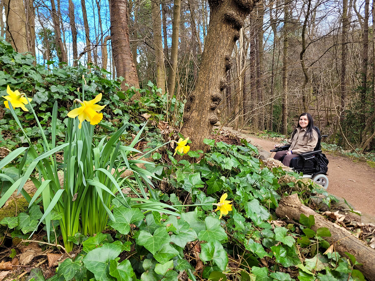 Emma enjoying a wheelchair accessible walk at Letham Glen. She is in the forest and there are daffodils next to Emma.