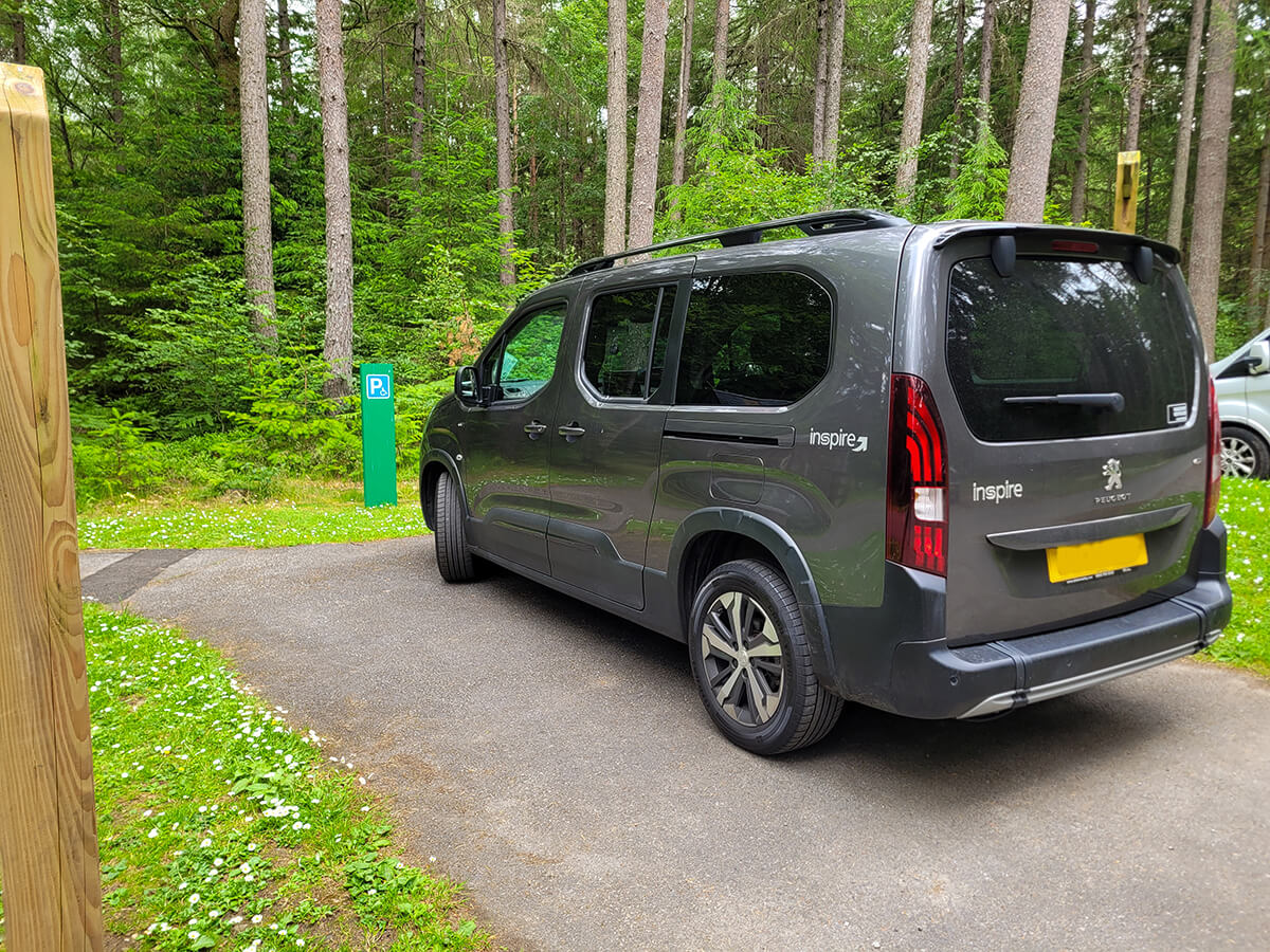 A grey wheelchair accessible vehicle parked in the car park of Faskally Woods. The car park is surrounded by tall pine trees.