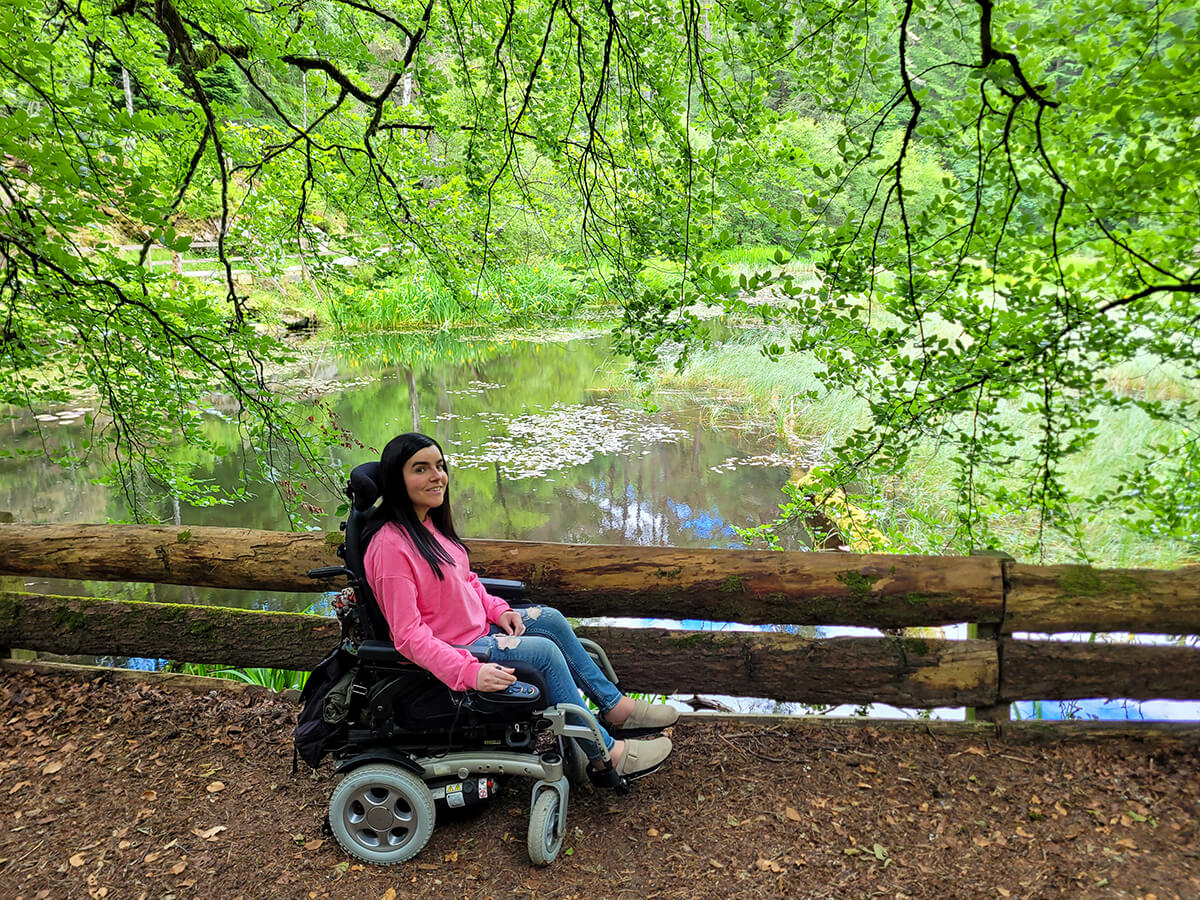 Emma on the wheelchair accessible path through the Dunmore Trail in Faskally Woods. Emma is wearing a pink sweatshirt, jeans and clogs. She has long straight black hair. She is sheltered by hanging trees and the loch is next to here. She is smiling at the camera.
