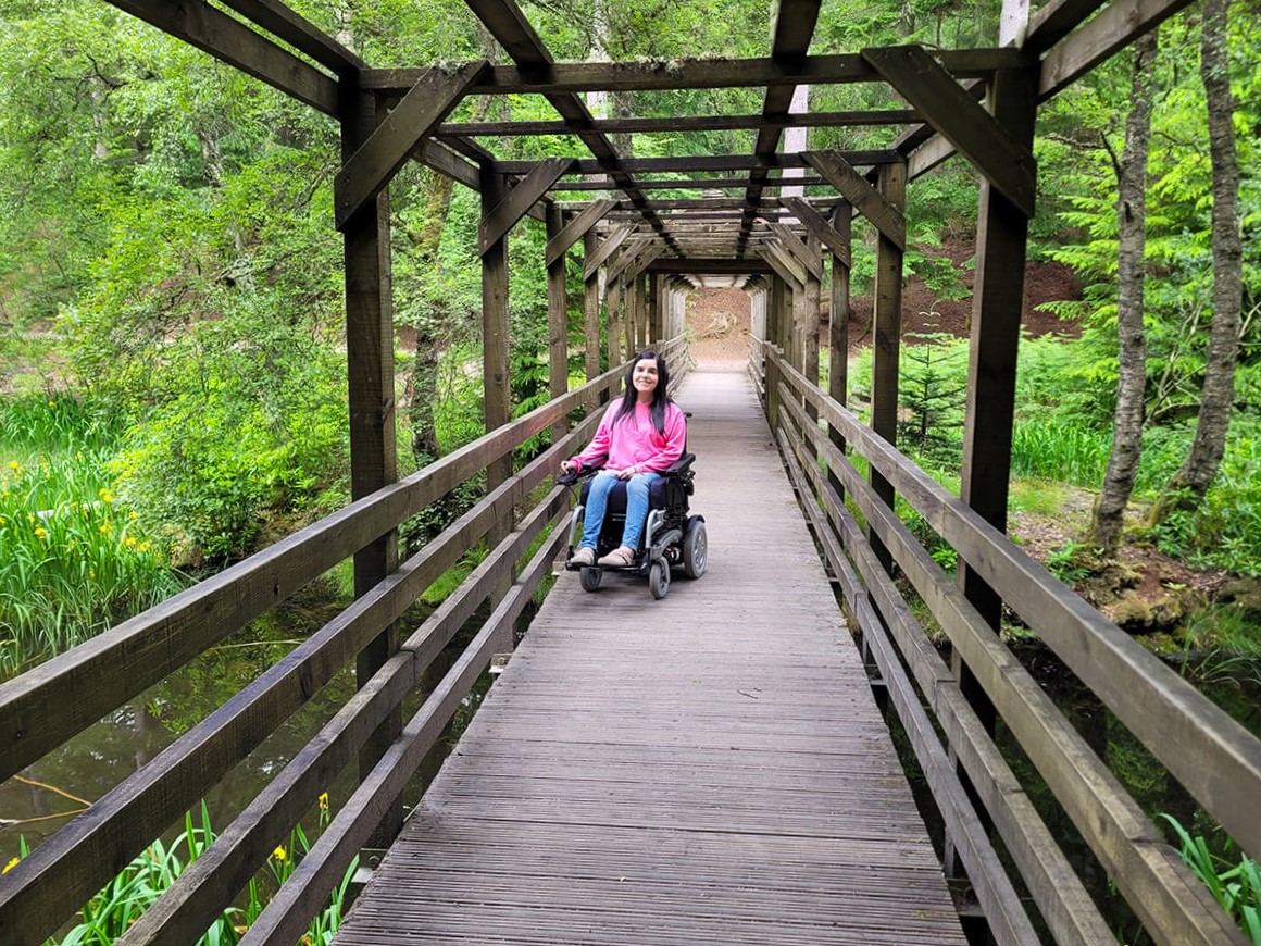 Emma on the level access wooden bridge through the Dunmore Trail in Faskally Woods. Emma is wearing a pink sweatshirt and jeans. She has long straight black hair. She is smiling at the camera.
