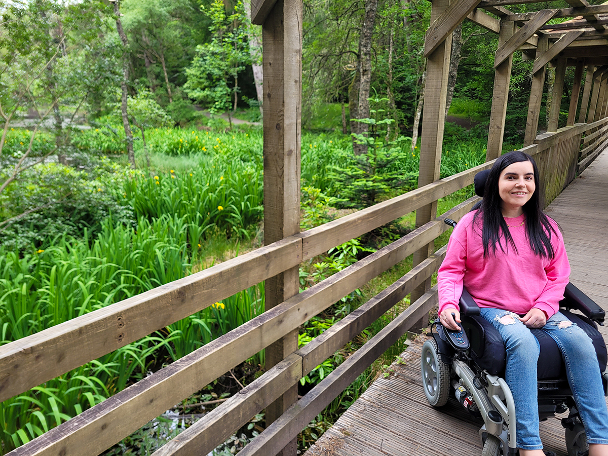 Emma on the level access wooden bridge through the Dunmore Trail in Faskally Woods. Emma is wearing a pink sweatshirt and jeans. She has long straight black hair. She is smiling at the camera.