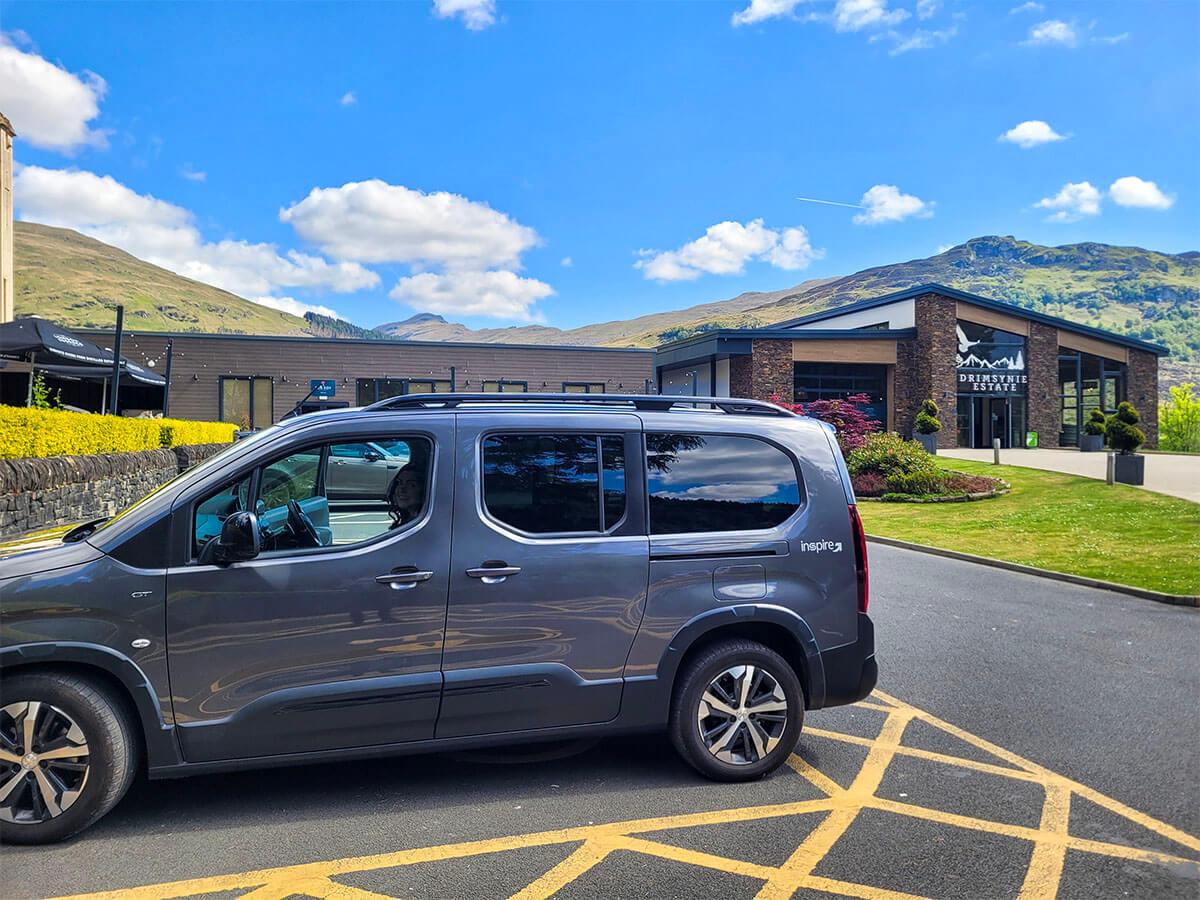 A grey wheelchair accessible vehicle parking in a disabled blue badge parking bay outside the Drimsynie Estate Hotel. The hotel has a beautiful backdrop of the Arrochar Alps