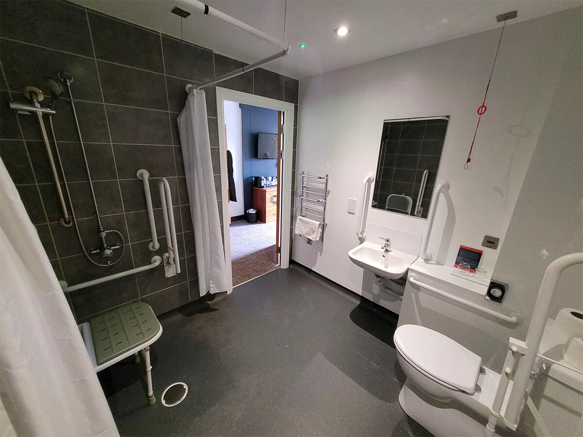 A white and grey decorated wetroom in the wheelchair accessible room at Drimsynie Holiday Village hotel. The photo shows the sink, toilet and roll in shower with shower seat.