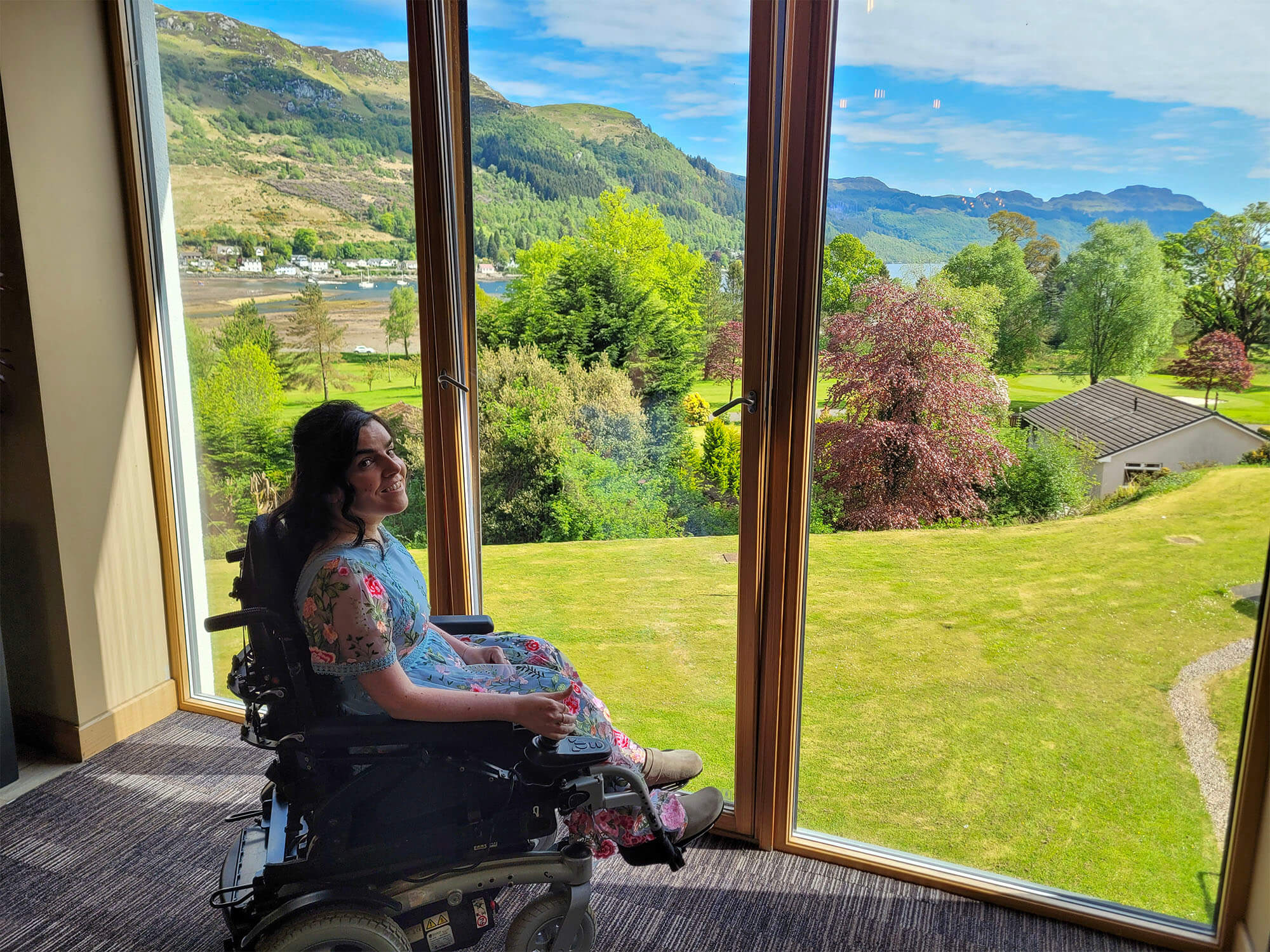 Emma is sitting in her wheelchair and is wearing a blue dress with pink embroidered florals. She is sat next to a floor-to-ceiling window in the Drimsynie Holiday Village hotel enjoying the beautiful view of the mountains and loch goil.