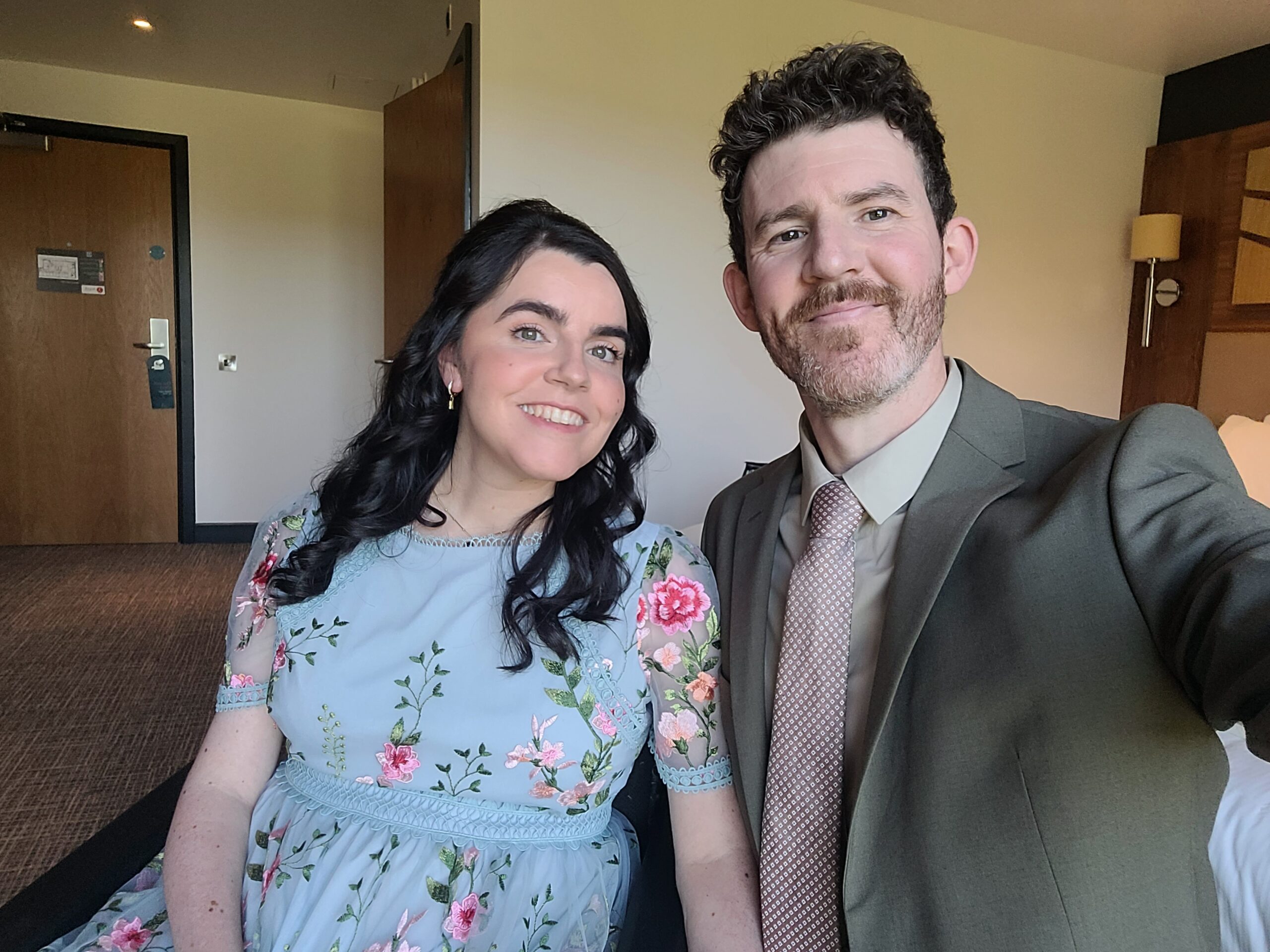 Emma is sitting in her wheelchair and is wearing a blue dress with pink embroidered florals. Standing next to Emma is her partner. He is wearing a light brown suit, shirt, and dusty pink tie. They are pictured in their hotel room at Drimsynie Holiday Village hotel before attending a family wedding in Lochgoilhead