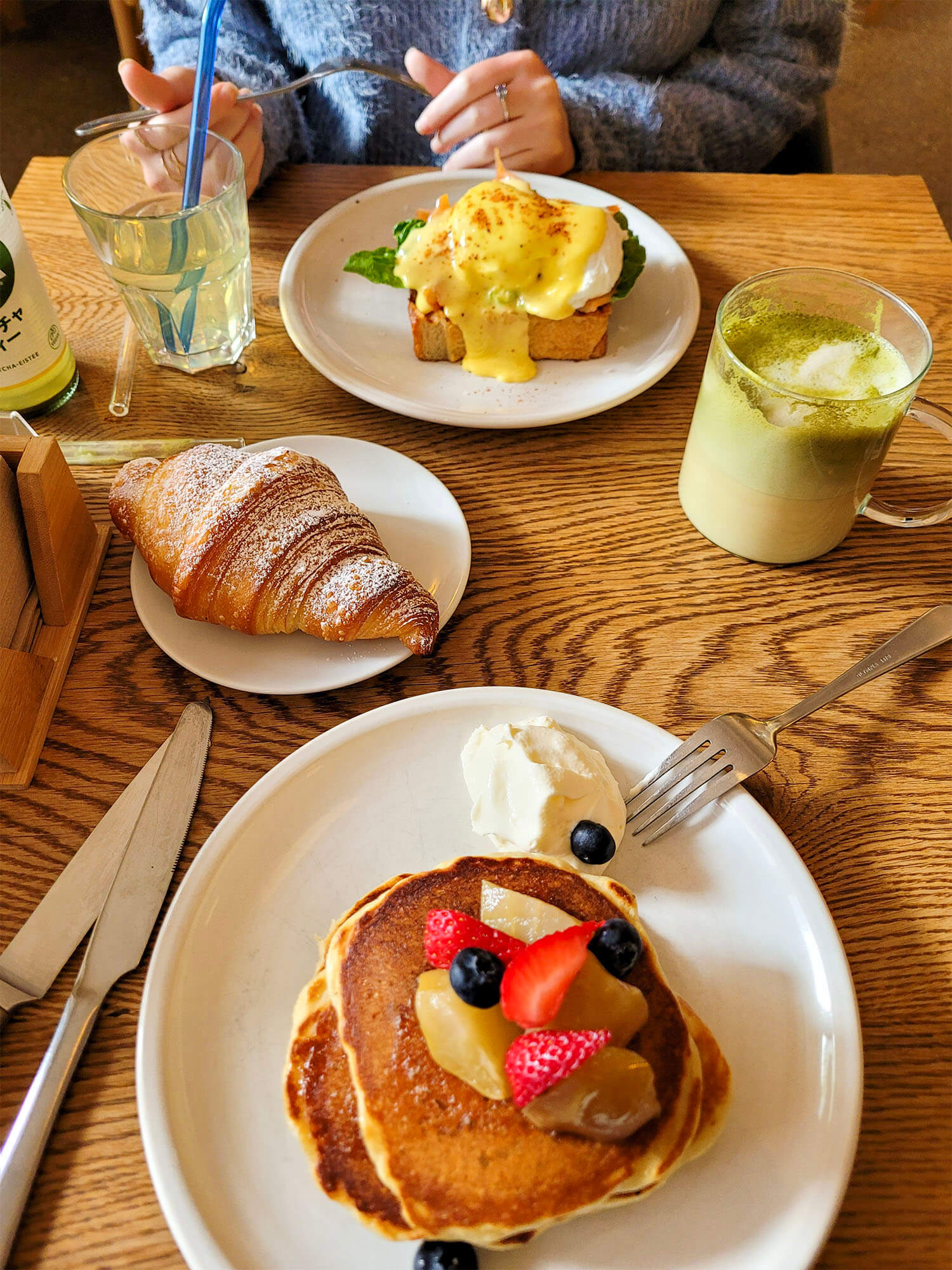 Eggs Benedict, a mug of matcha, a plate with a croissant and a plate with a stack of pancakes with fruit on top.