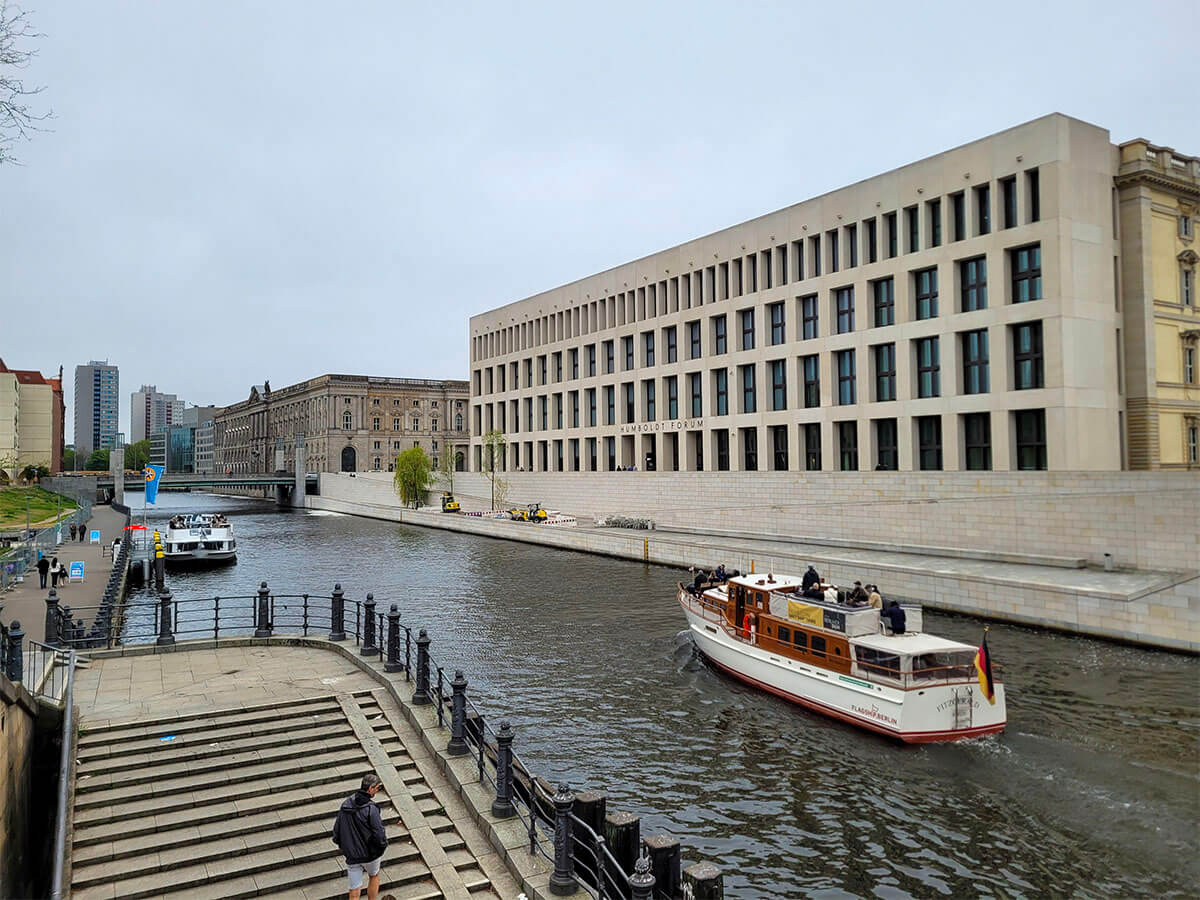 A shot of the Humboldt Forum on a dull day. A boat is sailing along the river in front of the building