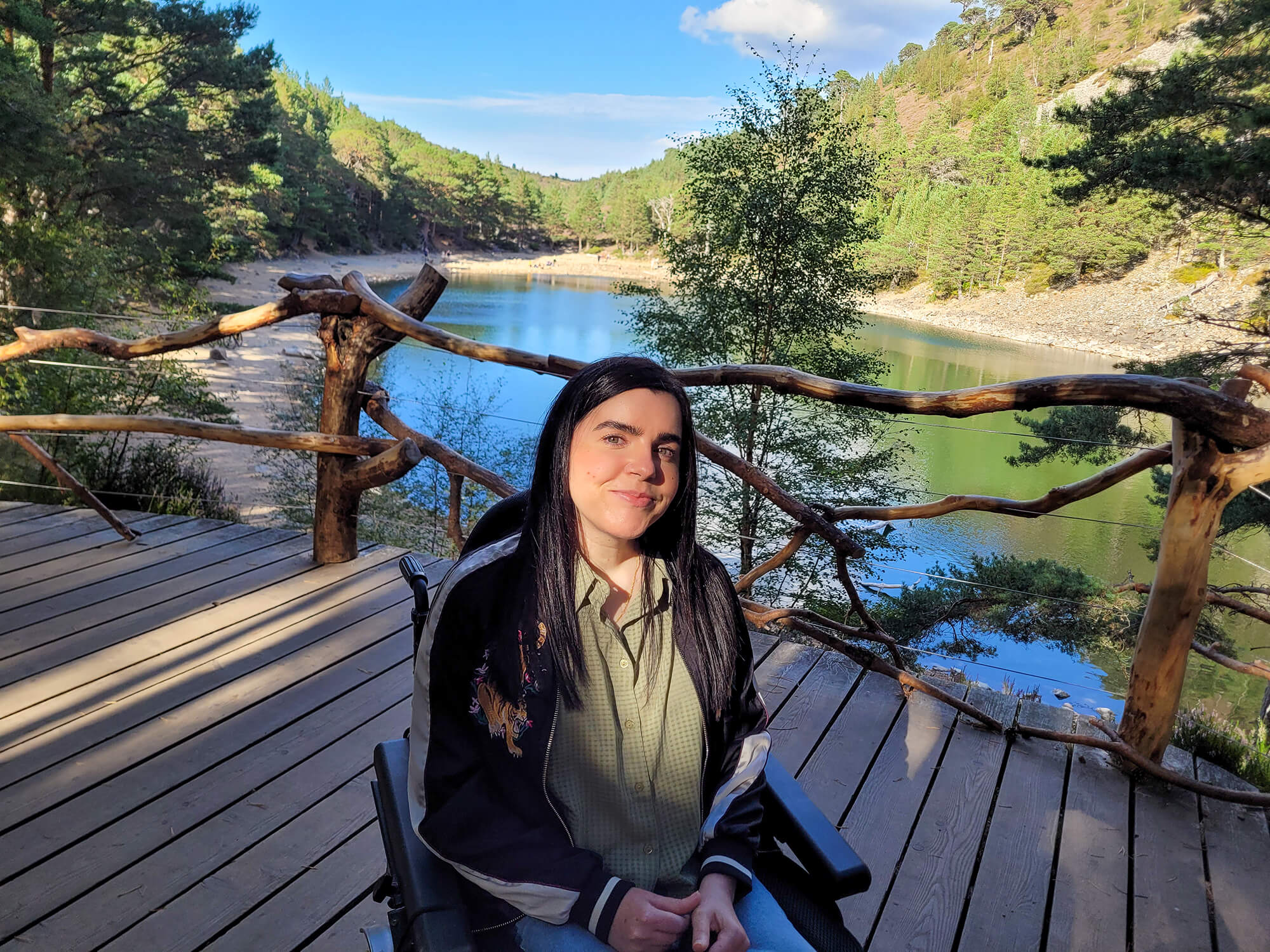Emma sat in her wheelchair on the viewing platform with an Lochan Uaine, the Green Loch behind her. She is smiling at the camera.