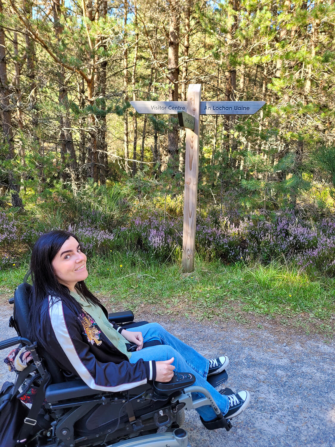 Emma sat in her wheelchair pictured next to a sign showing the direction of An Lochan Uaine. Emma is smiling at the camera.