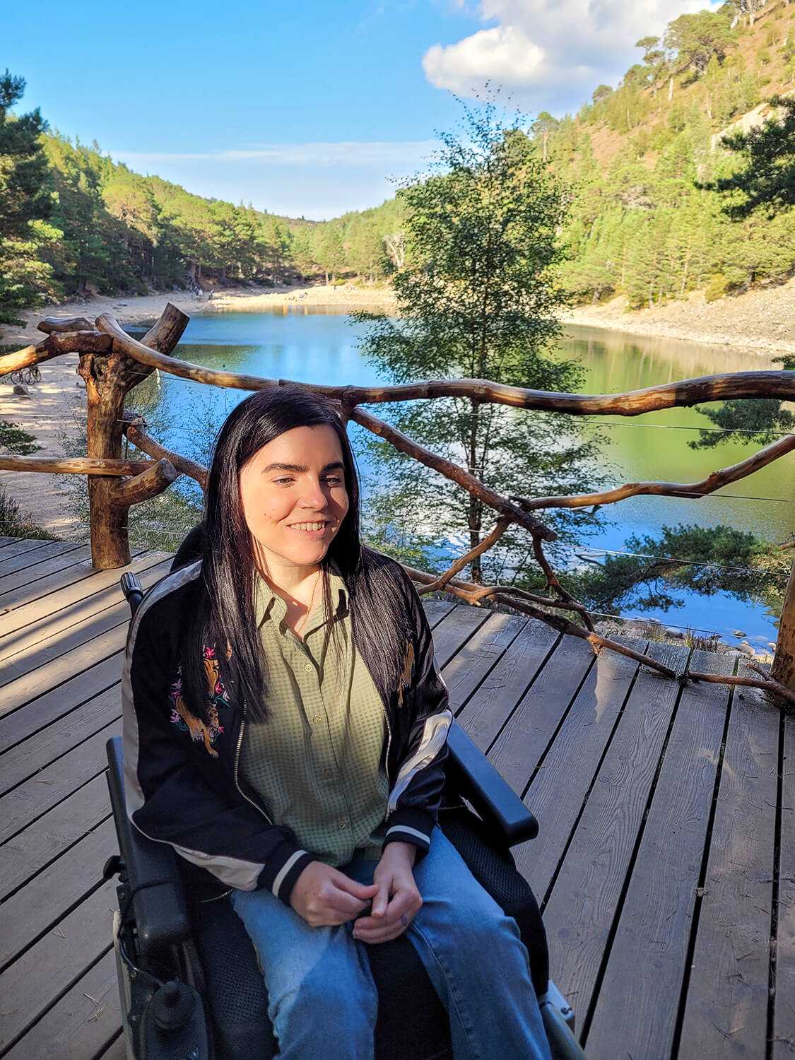 Emma sat in her wheelchair on the viewing platform with an Lochan Uaine, the Green Loch, behind her. She is slightly down towards the ground and smiling.
