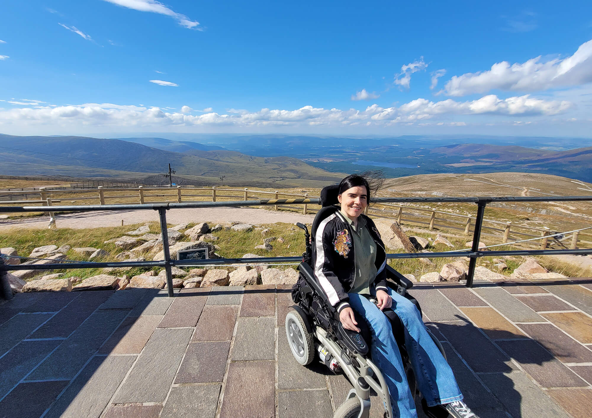 Emma sat in her wheelchair at the top of the Cairngorm Mountain Resort with a stunning view of the mountains and Loch Morlich in the background