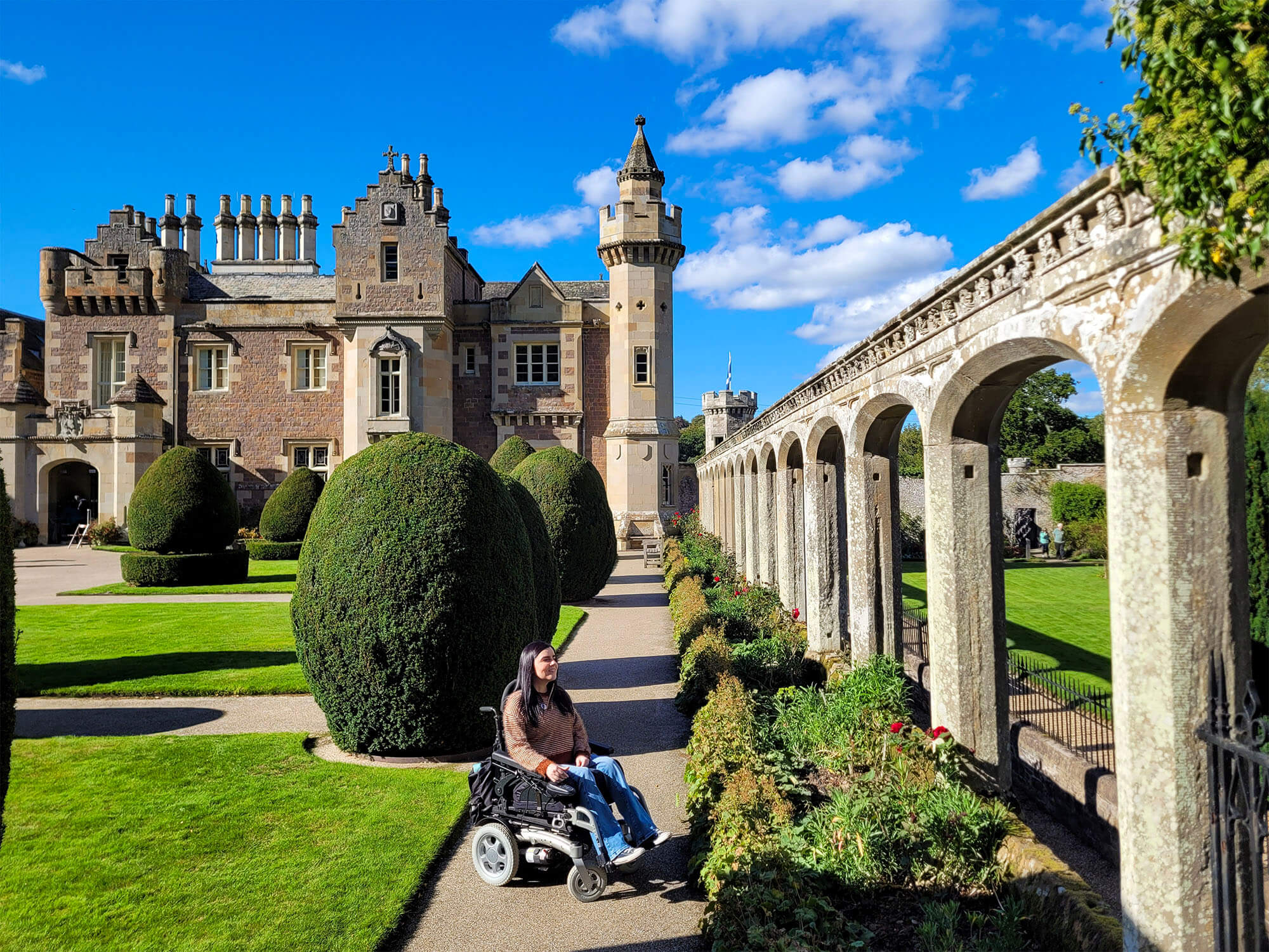 Emma, a power wheelchair user is in the beautiful manicured gardens with Abbotsford House behind her. Its a sunny day and Emma is looking up at the stone walled arches.