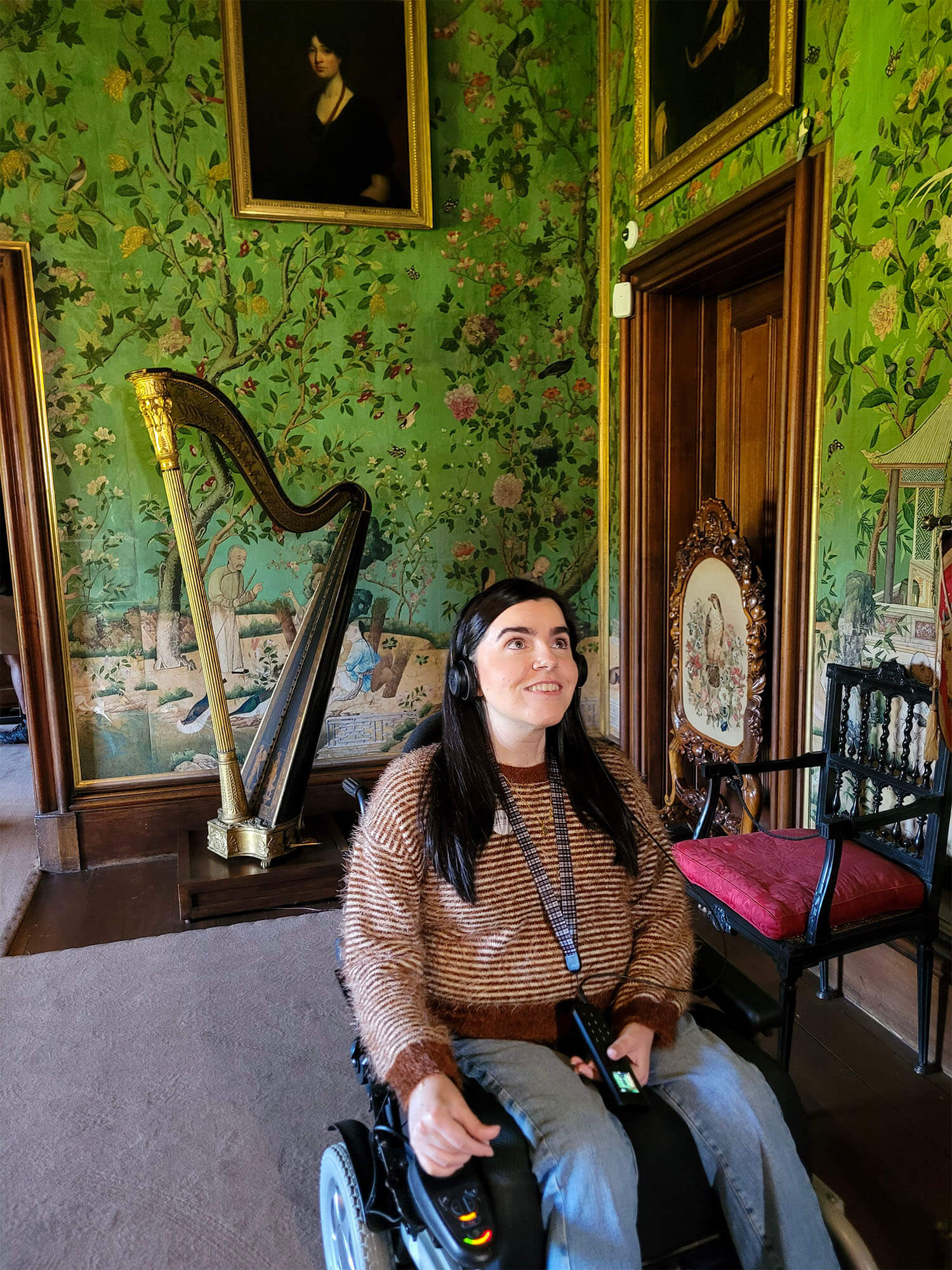 Emma, a power wheelchair user is wearing headphones for the audio tour and she is in the Chinese Room of Abbotsford House. She is looking up and smiling.