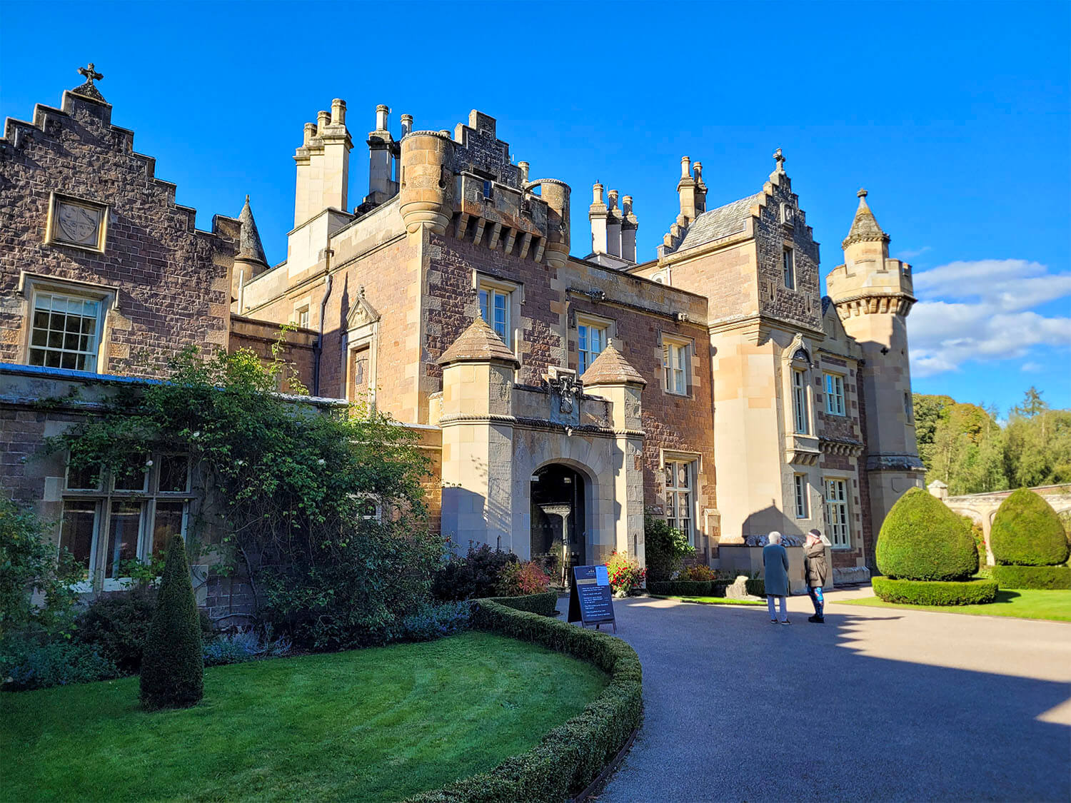 The exterior of Abbotsford House, the home of Sir Walter Scott, on a sunny day with blue sky. Two people are standing at the entrance.