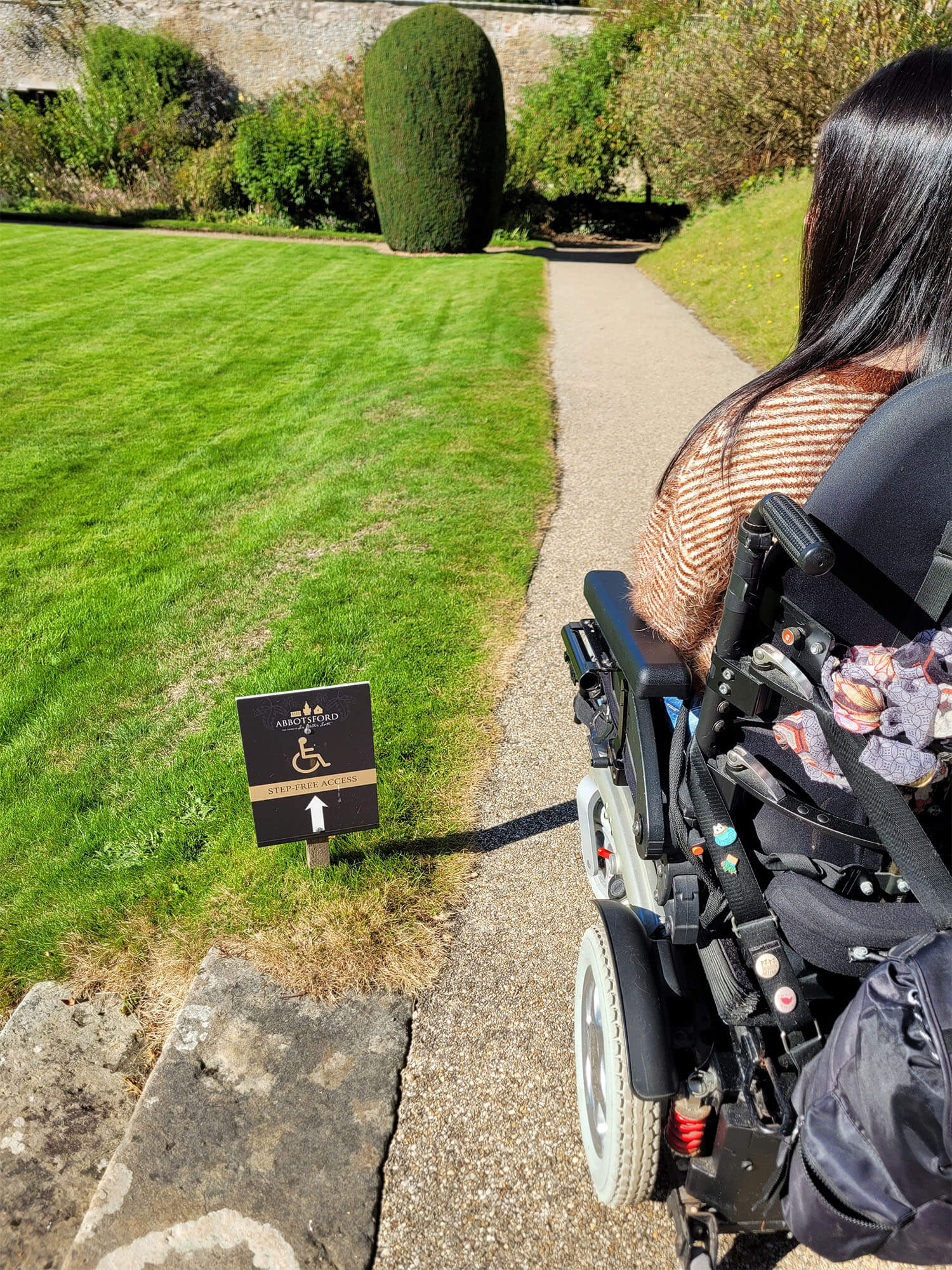 Emma, a power wheelchair user, is pictured in the beautiful garden at Abbotsford House next to a sign that reads. "step-free access"