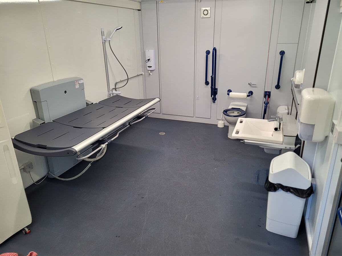 Interior of a Changing Places toilet showing the adult-sized changing bed, peninsular toilet and sink, ceiling-track hoist, grab rails and a shower.