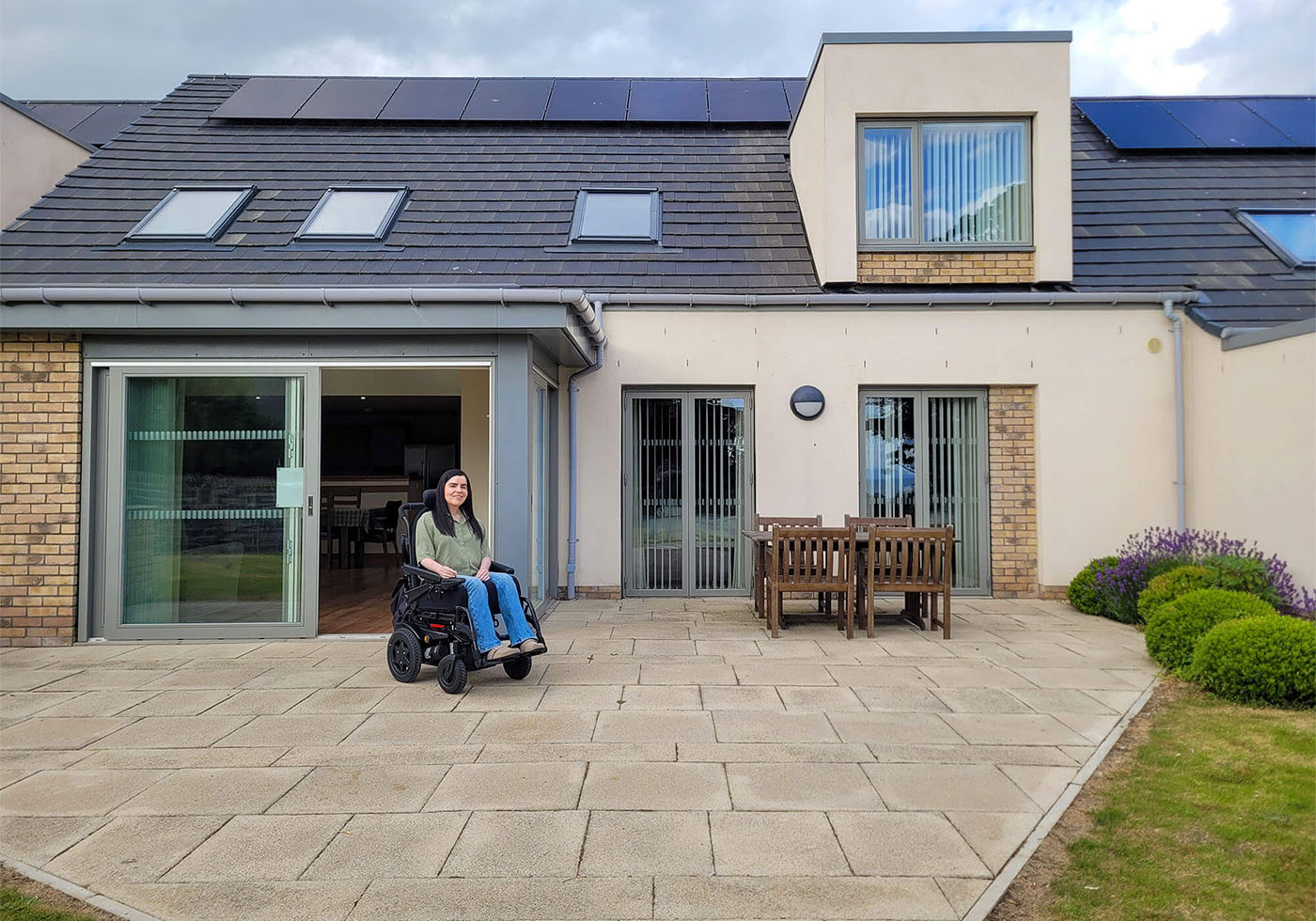 Emma in her wheelchair sat outside in front of the Largo lodge at Homelands Trust-Fife, a wheelchair accessible lodge in Scotland. Emma is wearing a green shirt and jeans.