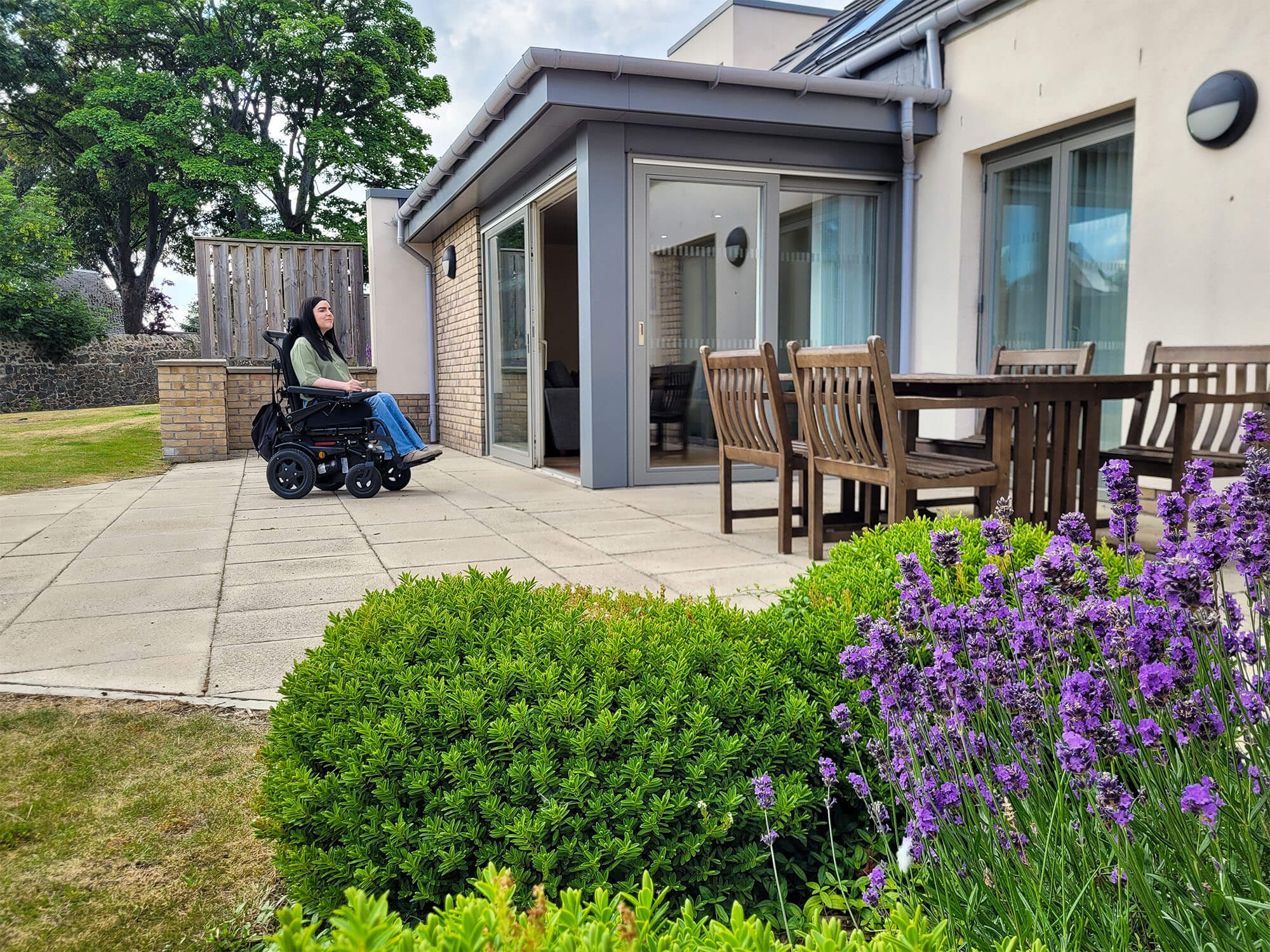Emma in her wheelchair sat outside in front of the Largo lodge at Homelands Trust-Fife, a wheelchair accessible lodge in Scotland. Emma is wearing a green shirt and jeans. Purple lavendar plants and green shrubs are in the fore front of the shot.